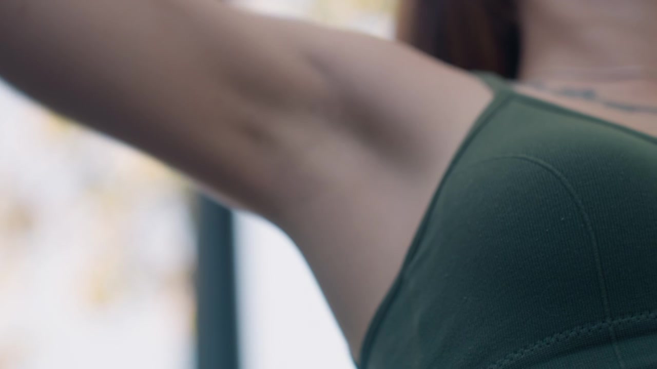 Primo piano di una giovane donna con lunghi capelli castani che indossa un top verde senza maniche, con il braccio alzato. La scena è illuminata delicatamente, al chiuso o con luce naturale, concentrandosi sui dettagli della texture della pelle.
