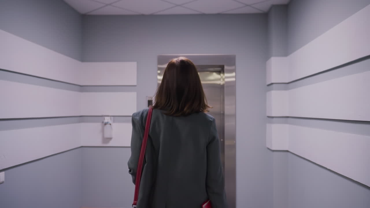 Businesswoman walking down modern office hallway toward elevator with briefcase, wearing formal attire in professional setting. Corporate environment, career, business concepts