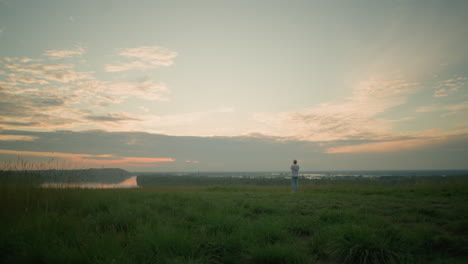 a man wearing a white shirt, hat, and jeans stands contemplatively in a grassy field by a tranquil lake at sunset. the serene landscape and warm evening light create a peaceful, reflective scene