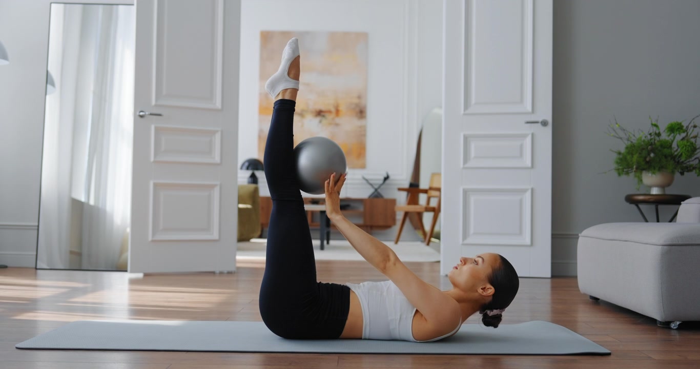 Giovane donna che esegue esercizi per il core con una piccola palla su tappetino da yoga a casa