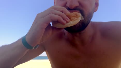 Bearded-man-eating-kebab-in-a-bun-on-the-beach-during-a-hot-summer-day