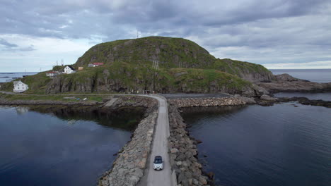 Aerial-drawback-dolly-shot-of-a-car-crossing-a-breakwater-wall-from-an-island-on-an-overcast-day-with-calm-seas