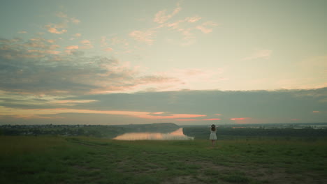 back view of a woman in a white dress and hat standing alone in an isolated grassy field by a tranquil lake at sunset. she gazes at the serene surroundings, embracing the peaceful moment of solitude