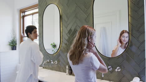 happy caucasian lesbian couple looking in mirror, improving hair and smiling in bathroom