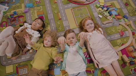 top view of four children lying on a carpet and waving at the camera raising arms in classroom in a montessori school