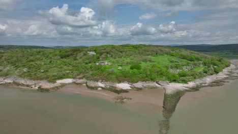 Verdant-rocky-coastline-with-houses-on-hillside-and-manmade-stone-structure-jutting-into-receding-tide-on-bright-day-in-Spring-at-Jenny-Brown's-Point,-Silverdale,-Lancashire,-England,-UK