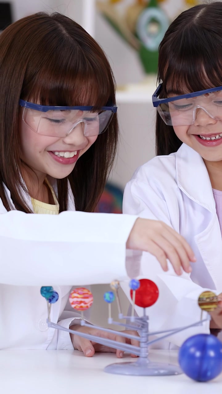 Two young Asian girls in safety goggles examine a planetary model in a science classroom.