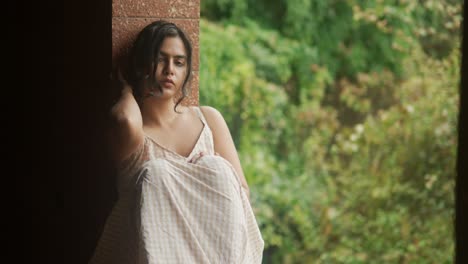 A-woman-in-a-white-dress-sits-relaxed-by-a-window-with-a-brick-wall-background,-gazing-outside