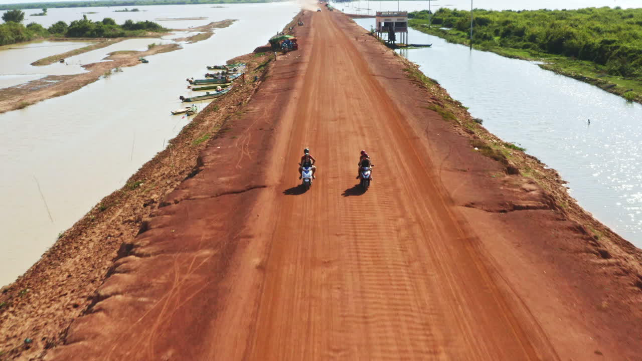 Aerial tracking of two motorcycles riding along flooded dirt rural Cambodian road near Kampong Phluk