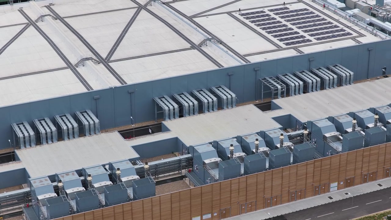 Large industrial server building featuring rooftop cooling systems and network infrastructure captured from high above during clear daylight hours in a suburban technology park