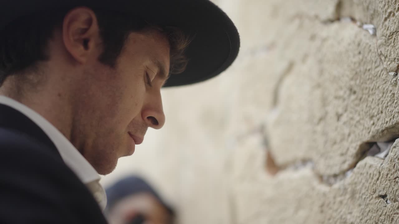 Jewish man praying at the Western Wall in Jerusalem. He wears a black hat and suit. Its a close up shot of his face from side. His eyes are closed as he prays he rocks backward and forwards.