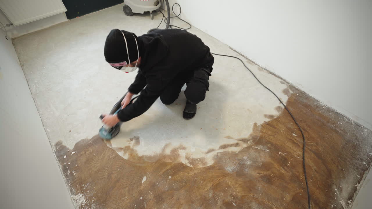 A person is sanding a floor surface using a power tool. The worker focuses on an area of the floor and removes old material. This is part of a renovation process in a room