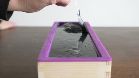 a women gently runs a spoon along a newly made block of black soap creating an artist pattern throughout the mould smooth surface of the soap