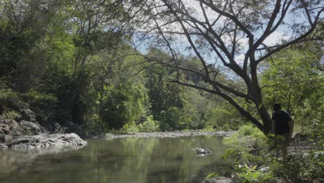 Tourist-dressed-in-a-cap-and-backpack-hiking-during-the-spring,-walking-along-the-rocky-bank-of-a-crystalline-river-in-the-department-of-El-Paraíso-in-Honduras