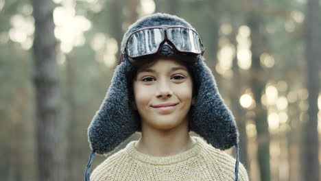 vista de cerca del pequeño niño caucásico soñador de ser piloto con sombrero y gafas especiales sonriendo a la cámara en el bosque