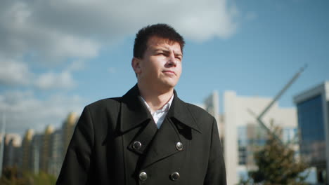 man in black coat strolling confidently during sunny day, surrounded by urban architecture with blurred buildings and trees in background under vibrant blue sky