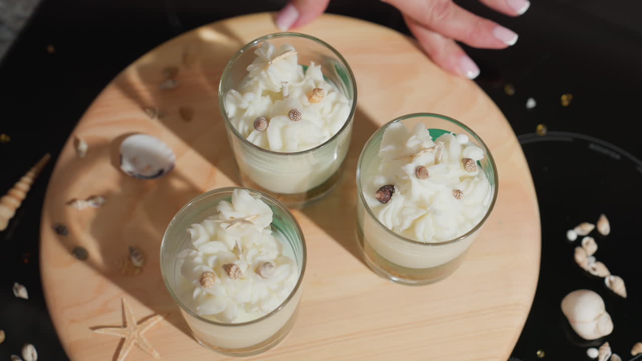 Culinary artist gently rotates wooden tray using fingertips to showcase three glass cups styled with milk-like whipped toppings and tiny shell decorations, forming elegant candle art in cozy kitchen