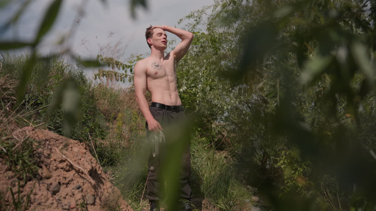 framed by leafy branches capturing warrior standing beside riverbank dirt path adjusting headband and stretching arms overhead in pose for intense outdoor survival challenge and movement behind