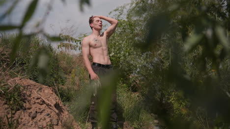 framed by leafy branches capturing warrior standing beside riverbank dirt path adjusting headband and stretching arms overhead in pose for intense outdoor survival challenge and movement behind