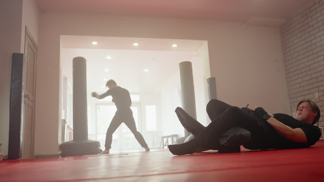 Athlete training in martial arts gym, one man practicing ground combat drills on dummy while another in background shadowboxes with gloves, showcasing power, stamina, discipline, concentration, and sports