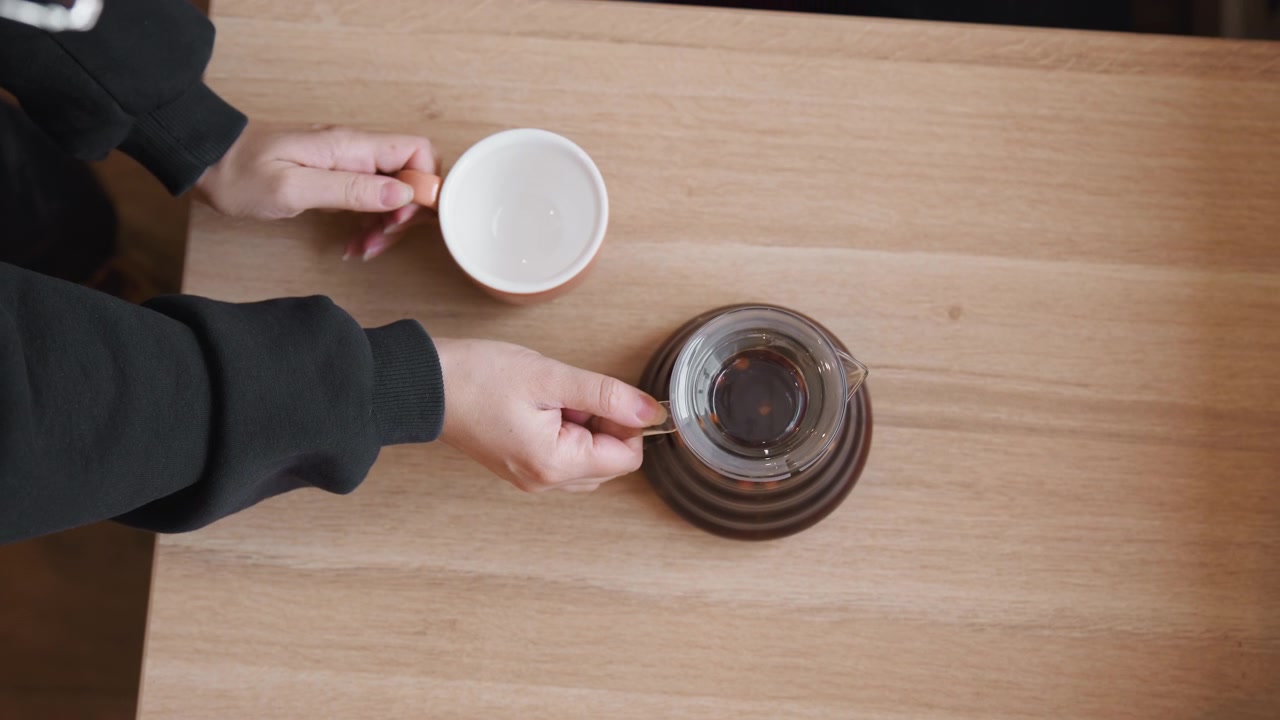 Primo piano di una persona che prepara il caffè aggiungendo un uovo in una caraffa di vetro su un tavolo di legno. La scena mostra le mani con oggetti come tazza, piattino e liquido trasparente in inquadrature stabili. Luce soffusa.