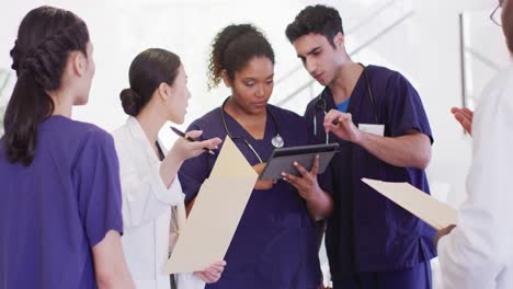 Video-of-diverse-group-of-male-and-female-doctors-looking-at-tablet-talking-in-hospital-corridor