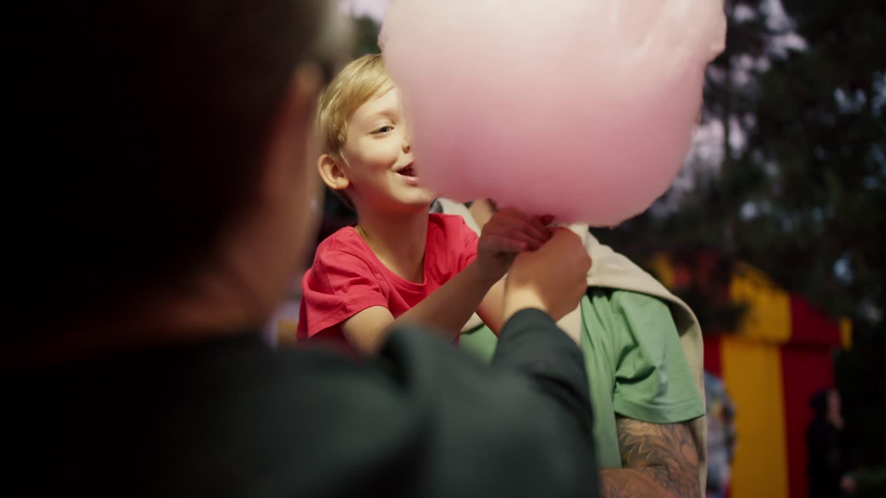 A little blond boy in a red T-shirt takes a large cotton candy from the hands of a cashier in an amusement park while sitting in the arms of his father in a Green T-shirt