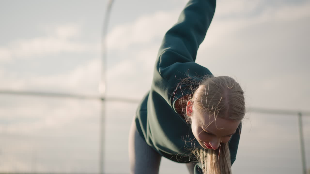 초록색 스웨터를 입은 젊은 여성이 피트니스 루틴에서 앞으로 구부러져 머리카락이 바람에 날아다니면서 에너지와 움직임을 보여주고 배경에는 명확한 야외 설정이 있습니다.