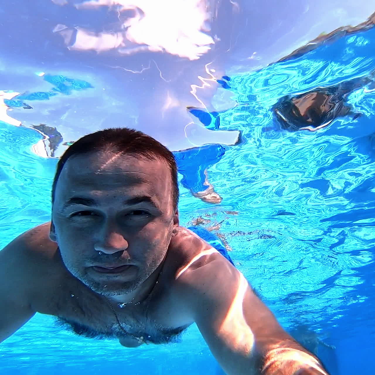 Swimmer swims forward while looking at camera underwater. Man swimming in clear water with underwater camera inside the swimming pool. Summer vacation.