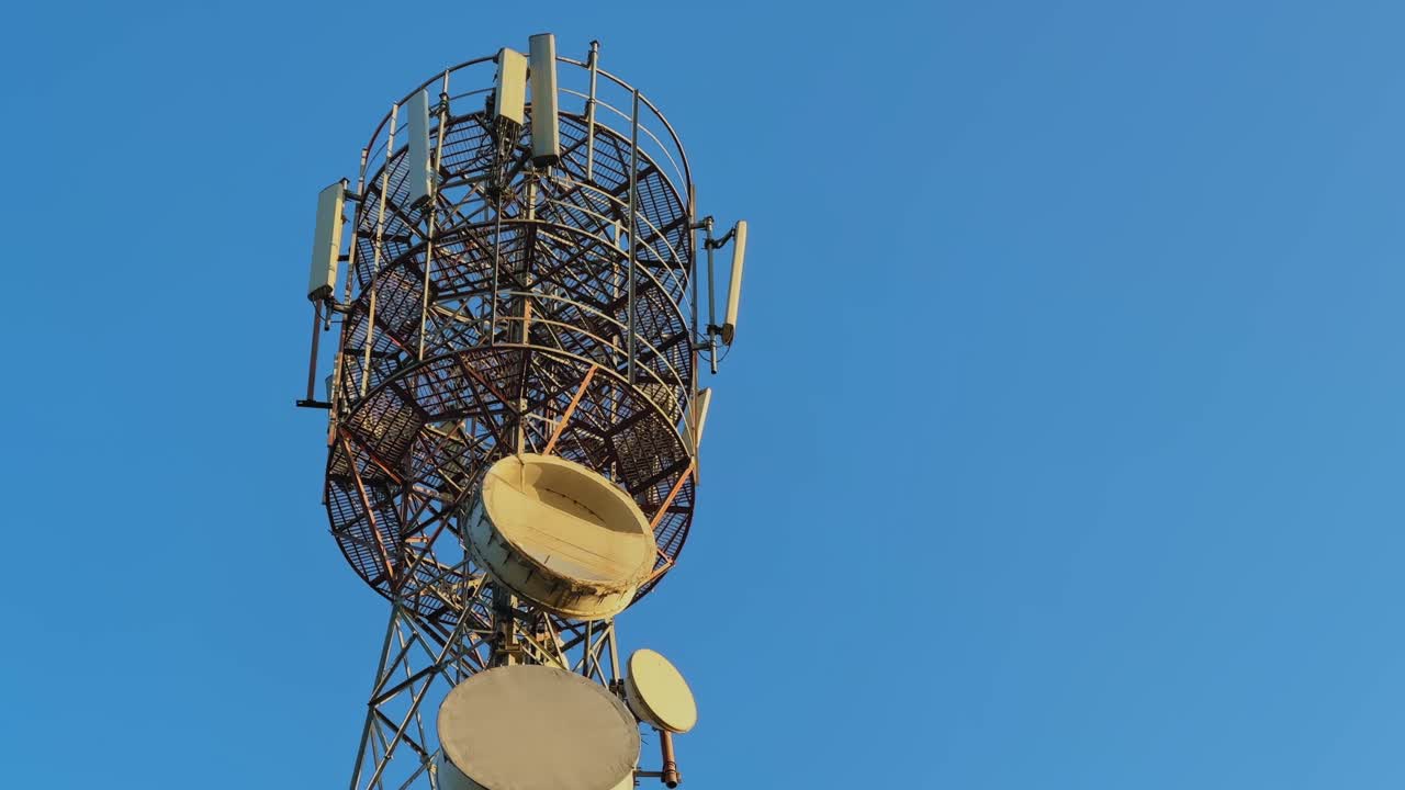 Closeup view of a tall telecommunication tower with circular metal framework, mounted antennas, and microwave dishes set against a clear blue sky, highlighting wireless communication infrastructure
