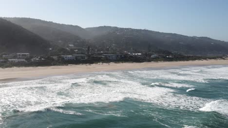 Las-Olas-Del-Océano-De-La-Costa-Salvaje-De-Sudáfrica-Se-Estrellan-En-Una-Larga-Playa-De-Arena-Con-Lujosas-Casas-De-Playa-En-El-Fondo