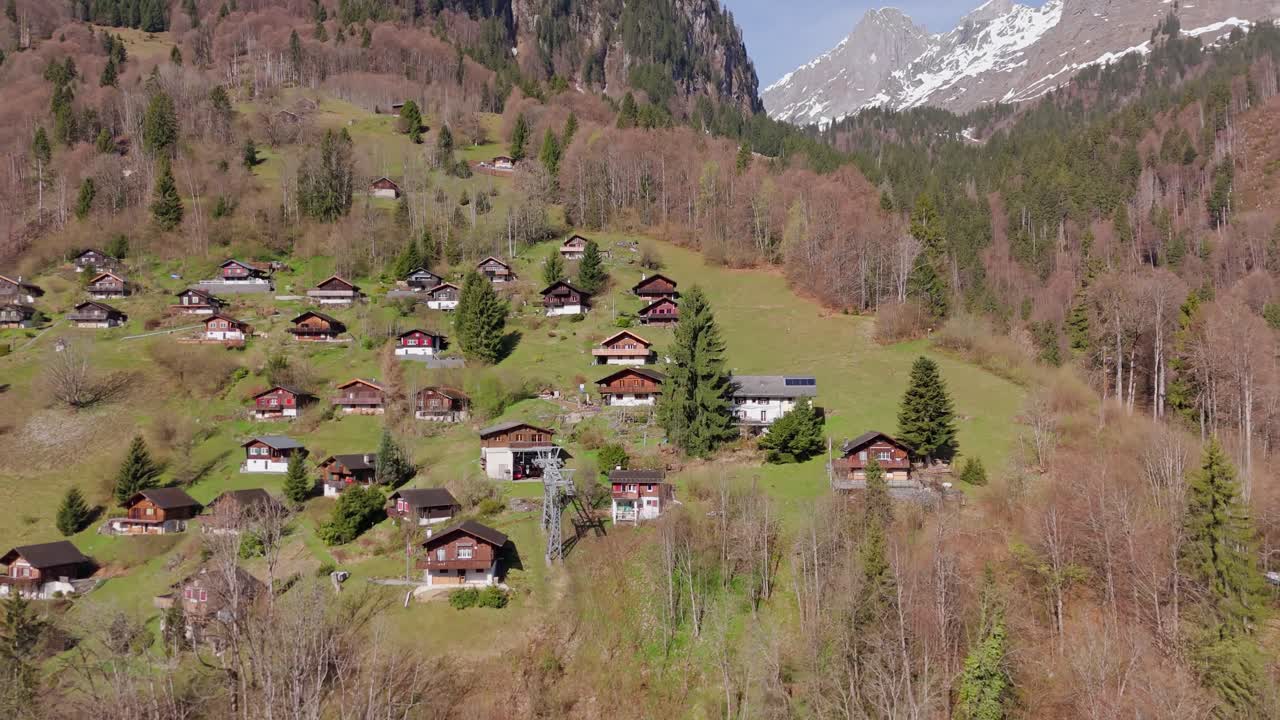 Alpejska osada Schlattberg nad Luchsingen w Szwajcarii z tradycyjnymi chatami na zboczu wzgórza i ośnieżonymi górami w tle. Odjazd kamery z powietrza.