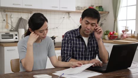 frustrated asian couple managing finances for the tax season with laptop in the dining room at home. they prop head feeling worried about financial problems while looking at document from bank