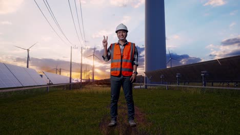 full body of asian male engineer with safety helmet smiling and showing peace gesture while standing with solar panel and wind turbines