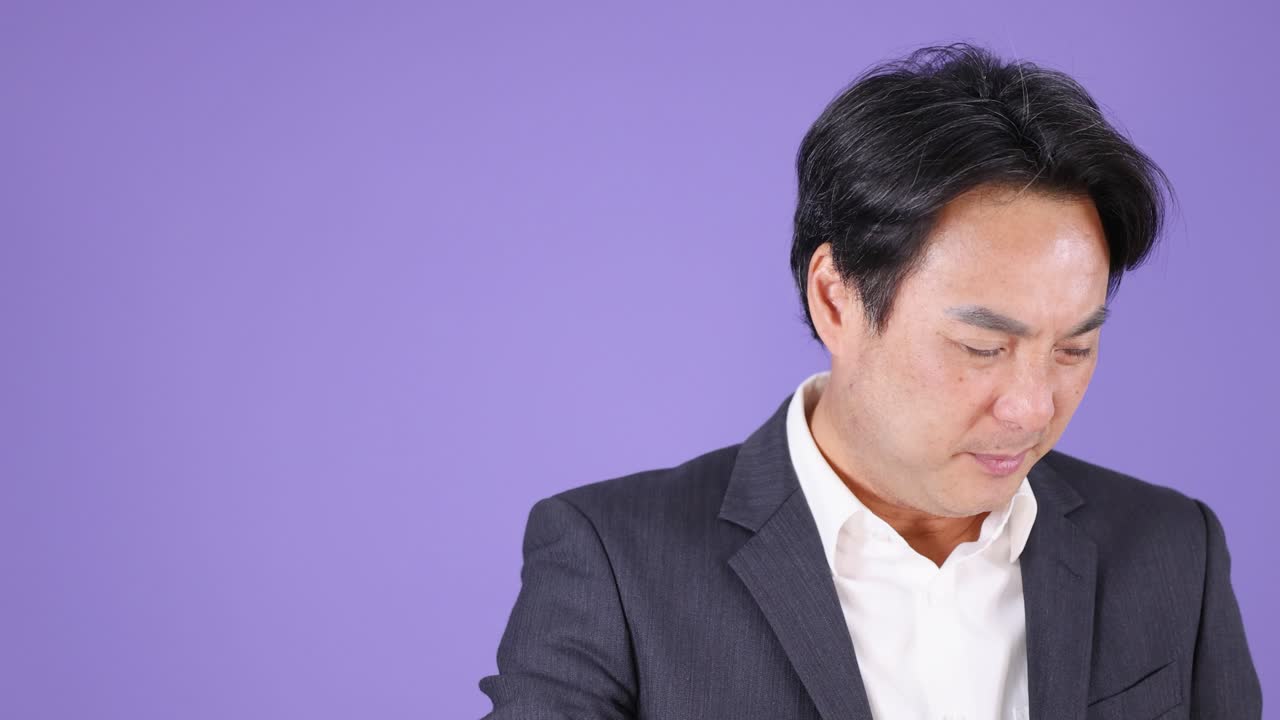 Asian businessman in formal suit touches neck, expressing discomfort, under bright studio lighting, static camera