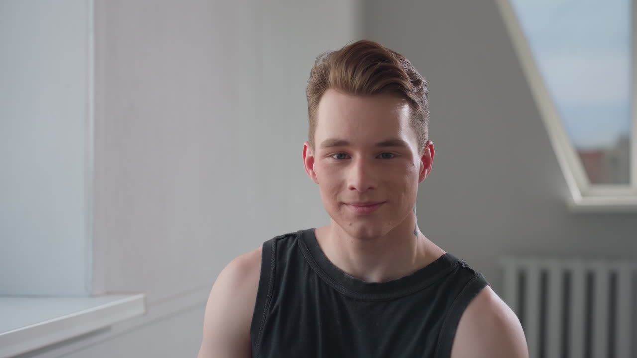 Young man in black sleeveless top sits by bright window in minimalist room, offers gentle smile while looking slightly down, relaxed posture conveys quiet confidence