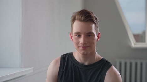 young man in black sleeveless top sits by bright window in minimalist room, offers gentle smile while looking slightly down, relaxed posture conveys quiet confidence