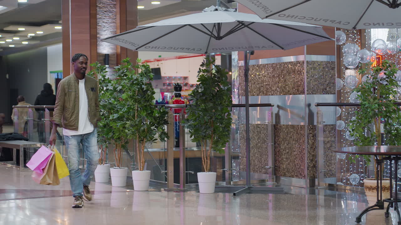 Young man holding colorful shopping bags walking inside modern shopping mall looking around with curiosity, wearing casual outfit of green jacket, white shirt and jeans near plants