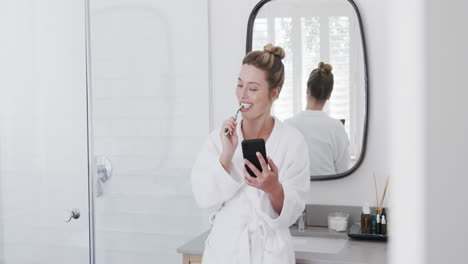 Happy-biracial-woman-brushing-teeth-using-smartphone-in-bathroom,-slow-motion