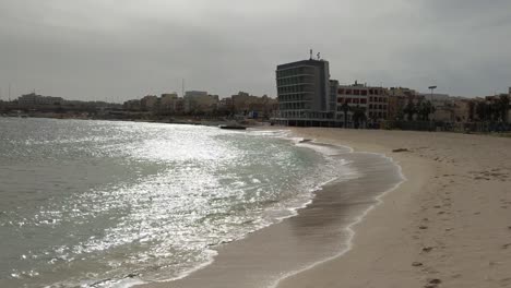 Calm Sea waves splashing in sand on sunny day with Water's Edge Hotel in background, Pretty Bay Coastline in Birzebbuga Malta