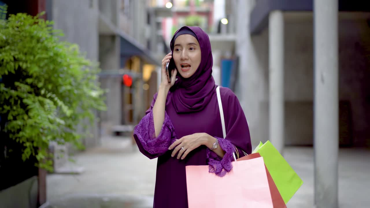 전통적인 헤드 스카프를 입은 젊은 무슬림 아시아 소녀가 거리를 고 전화로 이야기하고 가게에서 가방을 들고 있습니다. 여성 히자브는 쇼핑 후 드폰으로 이야기하고 웃고 있습니다.