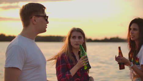 Two-female-students-are-moving-their-young-bodies-and-drinking-beer-on-the-hot-summer-party-on-the-beach-with-their-friend.