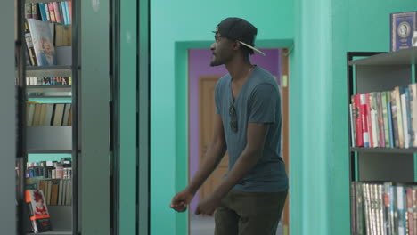 Young man in casual clothes dances joyfully in library hallway, wearing cap backwards and sunglasses hanging from shirt, moving with playful energy between bookshelves in bright modern interior