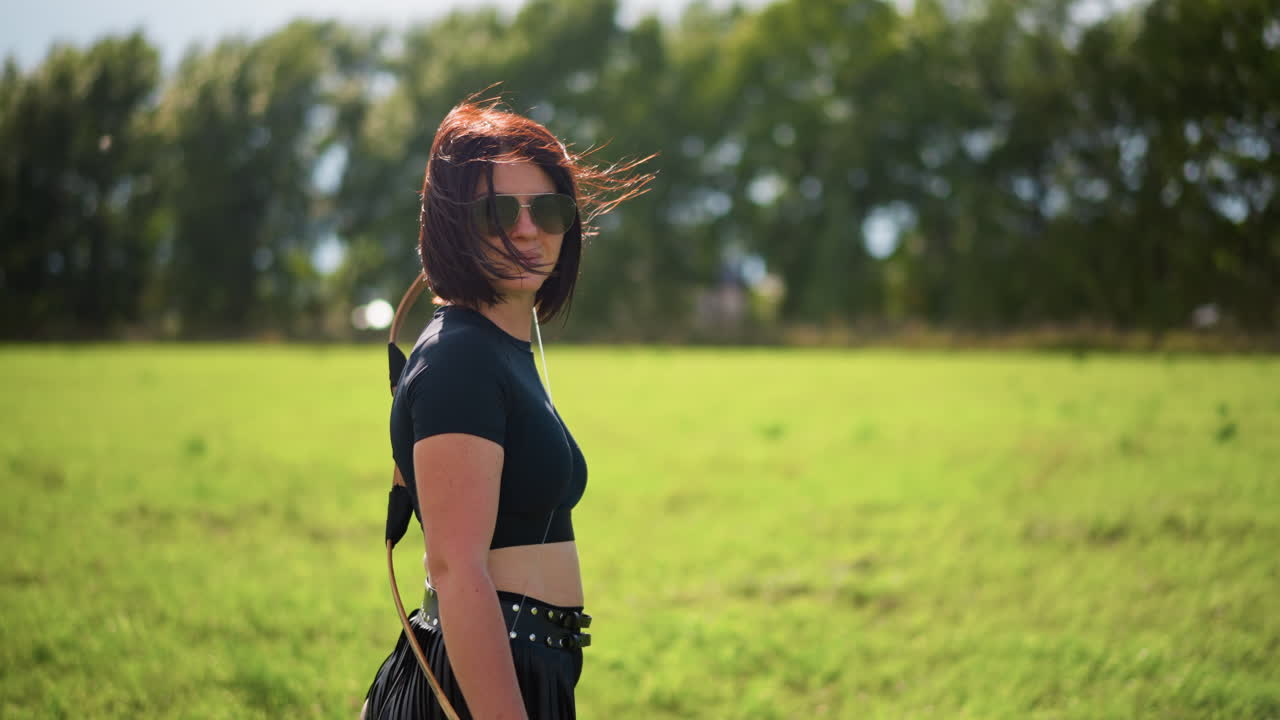 Arciera che cammina in un prato soleggiato, profilo laterale di una donna contemplativa che indossa un top corto nero e una gonna di pelle con frange borchiate e faretra sulla schiena, calda luce dorata, leggera brezza che increspa