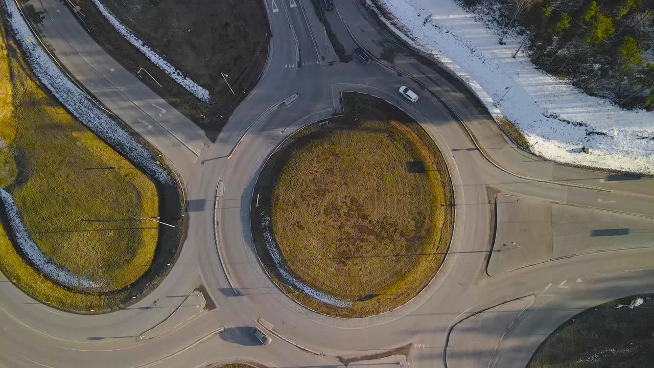 Top down aerial drone footage of a roundabout intersection in Laagri Saue district during a sunny autumn or spring day while cars, vehicles and trucks drive on the asphalt road. White snow visible.