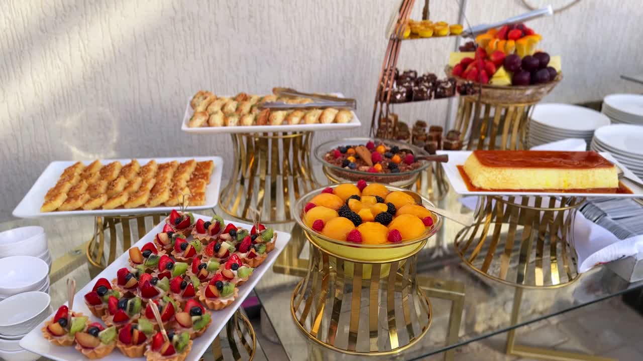 Primo piano 4K di un elegante buffet di dolci: mini tartellette alla frutta, involtini di baklava, cioccolatini, coppette di frutta e crème caramel su supporti dorati, ideale per matrimoni, hotel e catering