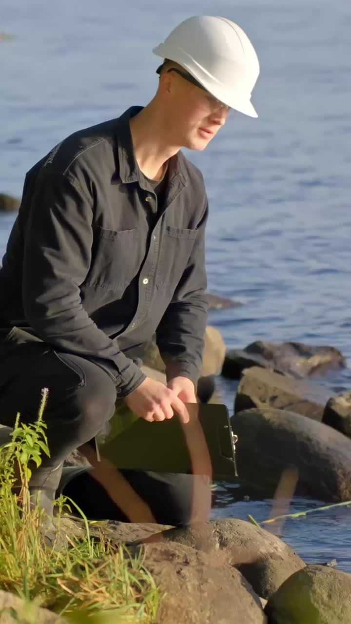 Fieldworker in outdoor gear leans and rubs rocks around side of the water, vertical