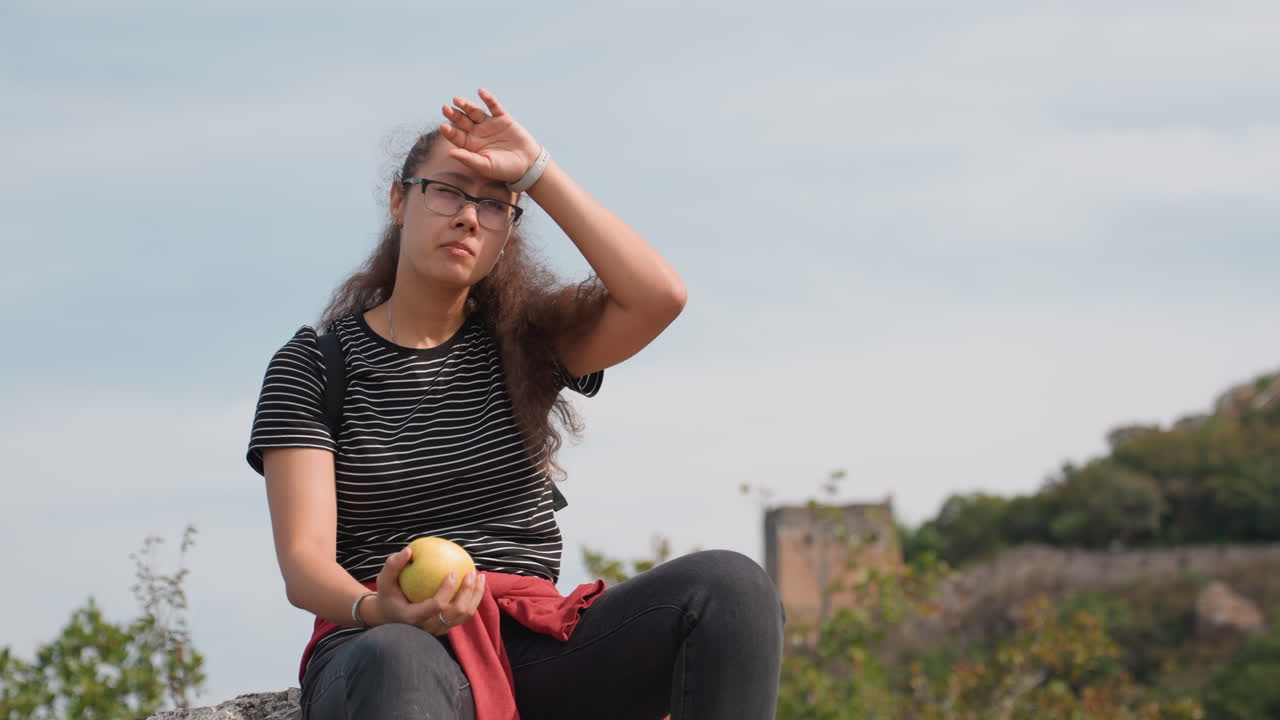 Joven sosteniendo una manzana en un mirador, dando un bocado y observando el cielo, top de rayas y gafas, chaqueta roja atada a la cintura, antiguo sendero de piedra detrás, vista desde la cima de la colina, una pausa de viaje casual, expresión curiosa.