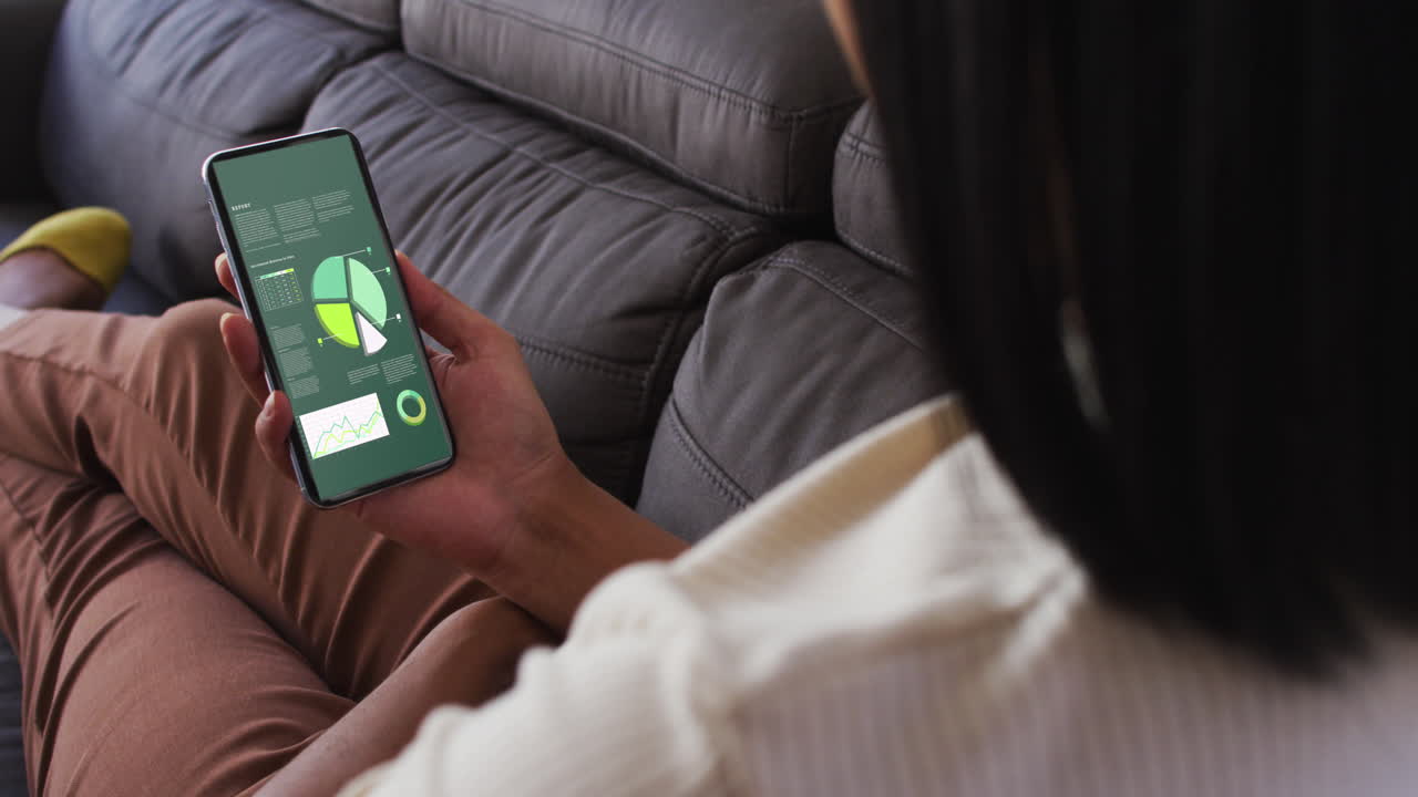 mujer biracial sentada en el sofá usando un teléfono inteligente con estadísticas en la pantalla