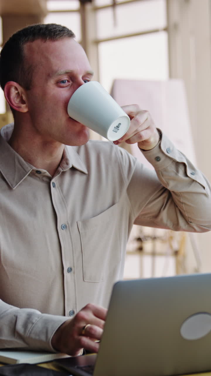 Resilient young man sitting at desk with laptop and having tea. Male having lunch break at workplace. Office backdrop. Vertical video